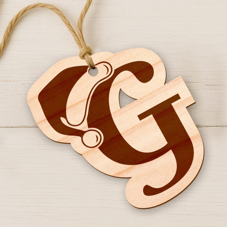 May include: Wooden ornament featuring the letters "CG" in a decorative font, with a dark brown stain on a light wood background. A natural twine loop is attached for hanging. The ornament is on a white wooden surface.