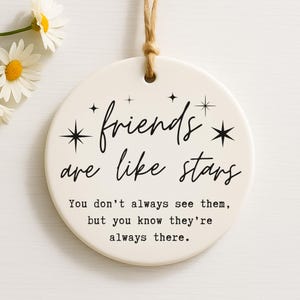 May include: A round, off-white ceramic ornament with the text "friends are like stars." The text is in black script, with star accents. The ornament has a jute string for hanging. Daisies are in the upper left corner.