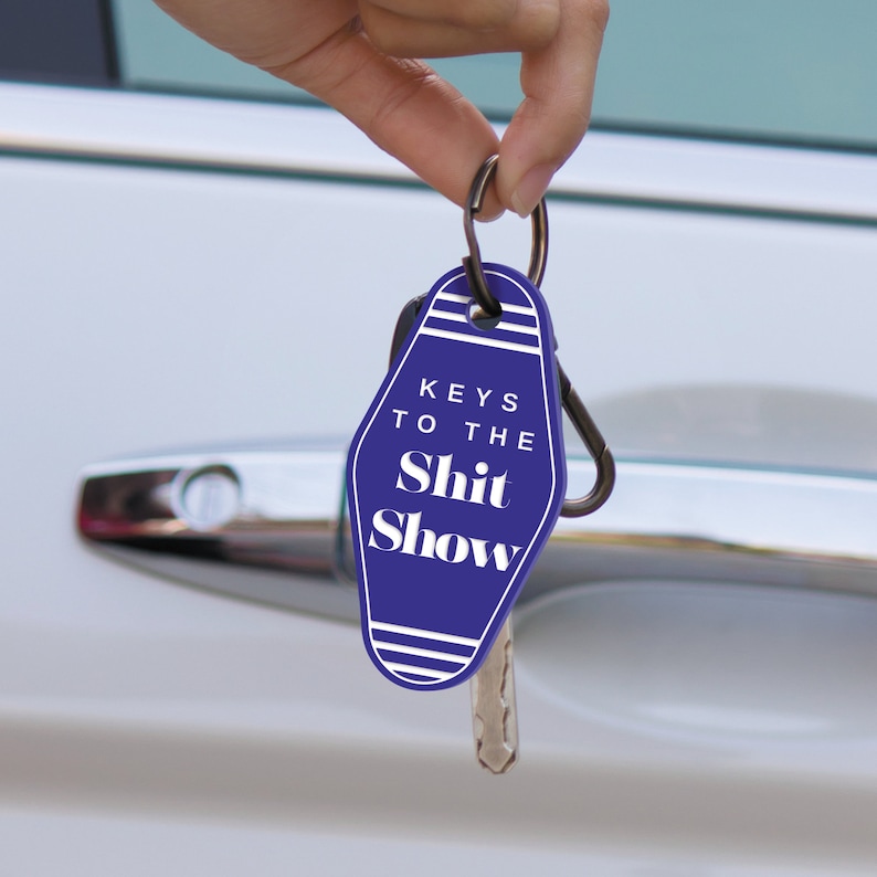 May include: A blue motel key chain with the text "Keys to the Shit Show" in white. The keychain is attached to a silver key ring and a silver carabiner.
