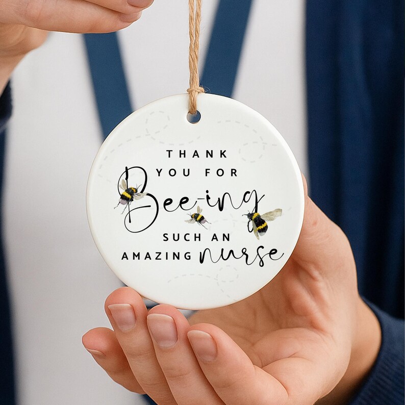 May include: A white ceramic ornament with the text "Thank You For Bee-ing Such An Amazing Nurse." The ornament features three illustrations of bees. A brown string is attached for hanging.