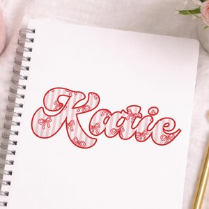 May include: A white spiral-bound notebook with the name "Katie" in red script. The letters are filled with a pink and white striped pattern and red bow illustrations. The notebook is on a white surface.