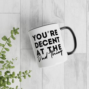 May include: White ceramic mug with a black rim and handle. The mug has black text that reads "You're Decent At The Dad Thing."