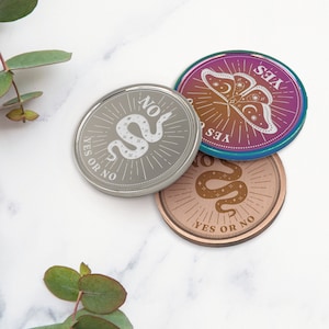 May include: Three round, metallic tokens with a yes or no theme. The first token is silver with a snake design and the text "YES OR NO". The second token is iridescent with a butterfly design and the text "YES". The third token is copper with a snake design and the text "YES OR NO".