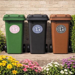 May include: Three wheeled trash bins in green, black, and brown. Each bin has a circular sticker with the number 35 in a decorative font. The bins are in front of a brick wall and a flower bed.