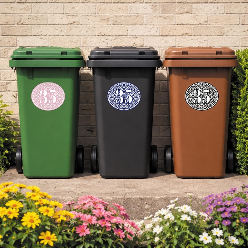 May include: Three wheeled trash bins in green, black, and brown. Each bin has a circular sticker with the number 35 in a decorative font. The bins are in front of a brick wall and a flower bed.