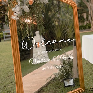 May include: A gold-framed mirror with the words "Welcome to our Wedding" in white script. The names "Mathew & Pheoby" and the date "26/04/2023" are also displayed. The mirror is decorated with flowers and greenery.