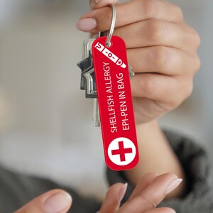 Op de afbeelding: Een rode sleutelhanger met de tekst "SHELLFISH ALLERGY EPI-PEN IN BAG" en een wit kruissymbool. De tag is bevestigd aan een set sleutels.