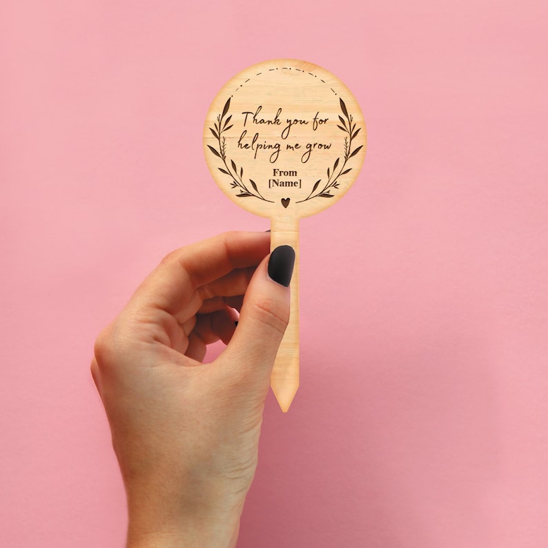 May include: Wooden garden stake with a round sign that says "Thank you for helping me grow. From [Name]" with a heart symbol. The stake is held in a hand.