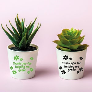 May include: Two white succulent pots with green plants. One pot has green flowers and the text "Thank you for helping me grow!" The other pot has black flowers and the same text.