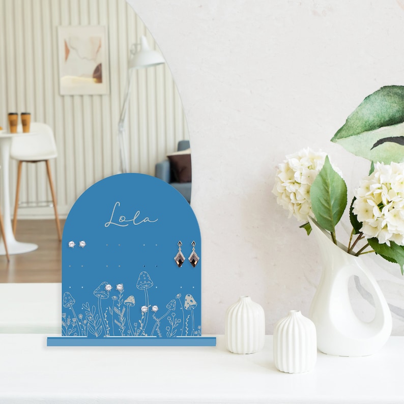 May include: Blue earring holder with a mushroom and floral design. The holder has the name "Lola" written on it.