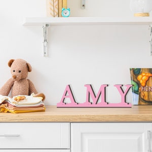 May include: Pink wooden letters spelling the name "AMY" on a wooden shelf. The letters have a 3D effect and are slightly raised.