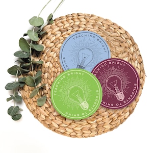 May include: Three round stickers with a lightbulb graphic and the text "Teaching Bright Students to Shine" in a circle around the lightbulb. The stickers are green, blue, and maroon and are on a woven straw mat.