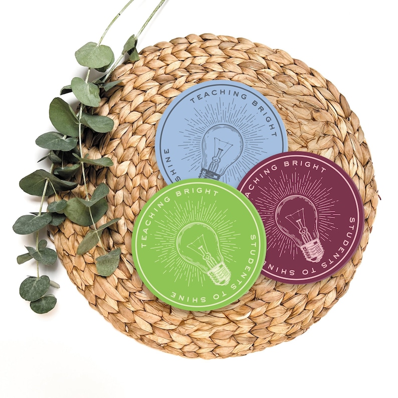 May include: Three round stickers with a lightbulb graphic and the text "Teaching Bright Students to Shine" in a circle around the lightbulb. The stickers are green, blue, and maroon and are on a woven straw mat.