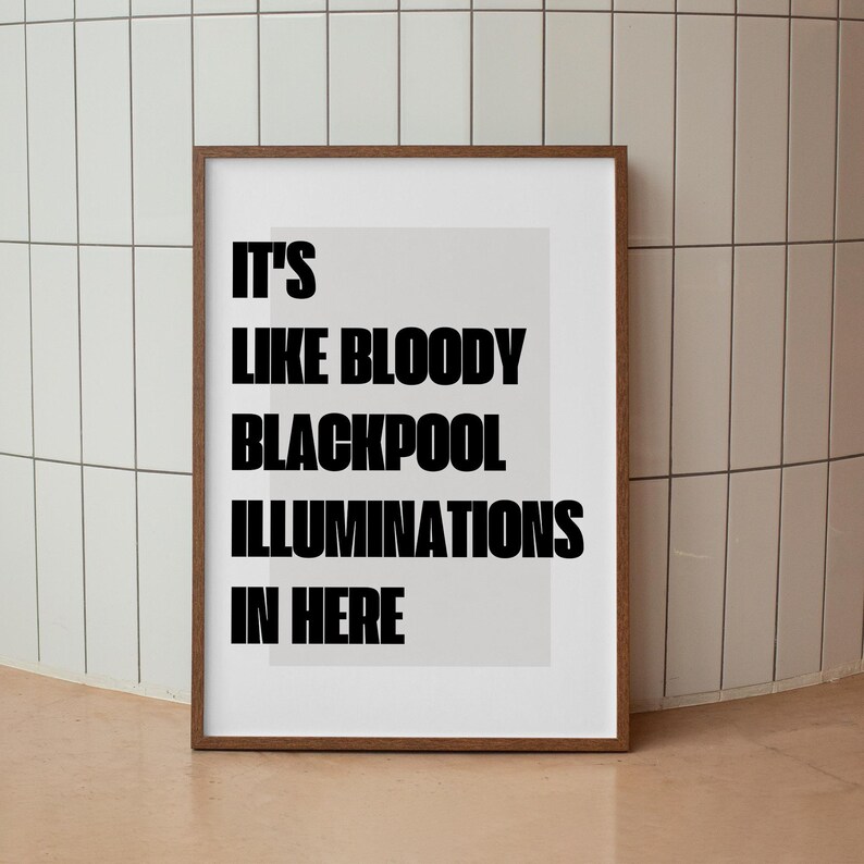 May include: A framed print with the text "IT'S LIKE BLOODY BLACKPOOL ILLUMINATIONS IN HERE" in bold black letters. The print has a light gray background and a brown wooden frame. The image is displayed against a white tiled wall.