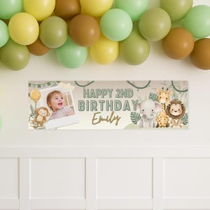 Puede incluir: Una pancarta de cumplea&ntilde;os con el texto "HAPPY 2ND BIRTHDAY Emily" y una foto de un ni&ntilde;o. La pancarta tambi&eacute;n presenta ilustraciones de un mono, un elefante, una jirafa y un le&oacute;n. Globos verdes, amarillos y marrones est&aacute;n arriba.