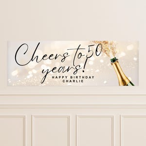 以下が含まれることがあります： 「Cheers to 50 years! Happy Birthday Charlie」と書かれたお祝いのバナー。シャンパンのボトルが飛び出し、金色の輝きが散っています。背景は、柔らかくぼやけた光のボケ効果があります。