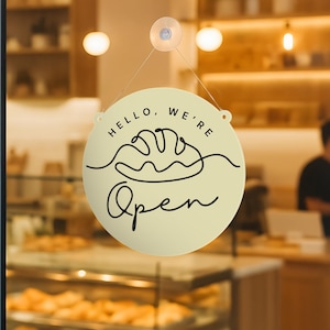以下が含まれることがあります： 「HELLO, WE'RE Open」の黒い筆記体とパンの線画が描かれた、丸いクリーム色の看板。お店の窓に吊り下げられており、営業中であることを示しています。