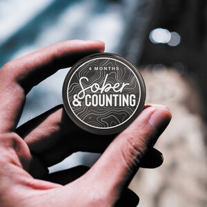 May include: A black and white round coin with the text "4 Months Sober & Counting" on a white background. The coin is being held in a person's hand.