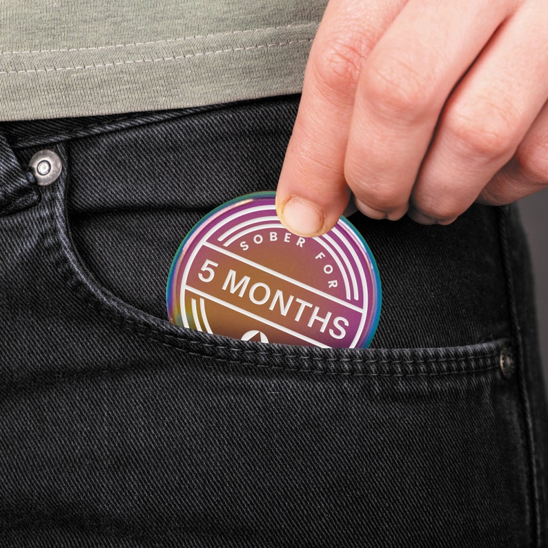 May include: A person's hand is holding a round, iridescent sobriety counter in a pocket. The counter has the text "SOBER FOR 5 MONTHS" printed on it.