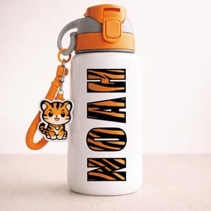 May include: White water bottle with orange accents and the name "NOAH" in tiger stripe lettering. Includes a matching orange strap and a tiger charm. The bottle has a gray handle and a push-button lid.
