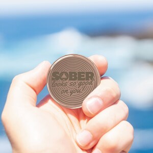 Pu&ograve; includere: Una moneta rotonda di colore rame con una superficie testurizzata e le parole "SOBER looks so good on you!" in lettere marroni.