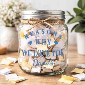 May include: A clear glass jar filled with folded notes, tied with twine, and a silver lid. The jar has blue text that reads "REASONS WHY WE LOVE YOU Daddy" with blue heart icons. Scattered notes are on the wooden surface.