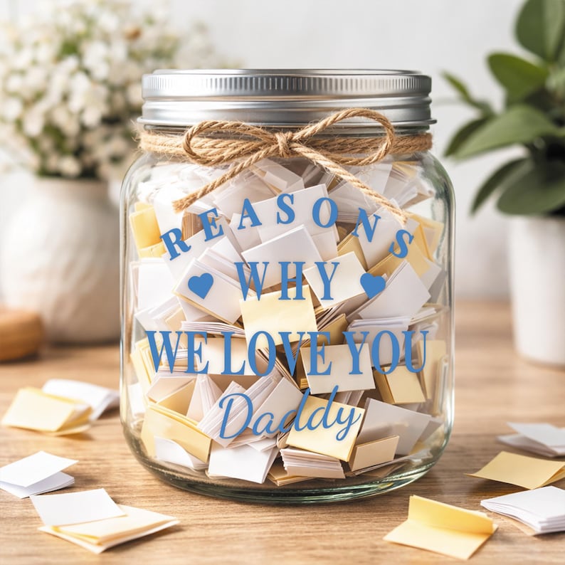 May include: A clear glass jar filled with folded notes, tied with twine, and a silver lid. The jar has blue text that reads "REASONS WHY WE LOVE YOU Daddy" with blue heart icons. Scattered notes are on the wooden surface.