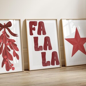 May include: Three framed art prints with a holiday theme. The prints feature a red mistletoe with a bow, the words "FA LA LA" in red, and a red star. The prints are in light-colored wooden frames and are displayed on a light-colored wooden floor.