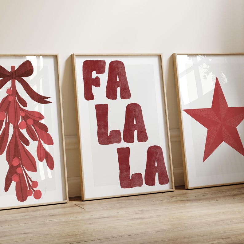 May include: Three framed art prints with a holiday theme. The prints feature a red mistletoe with a bow, the words "FA LA LA" in red, and a red star. The prints are in light-colored wooden frames and are displayed on a light-colored wooden floor.