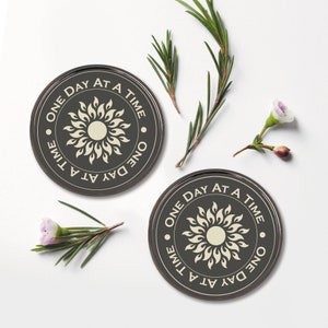 May include: Two black round coasters with a white sun design and the text "One Day At A Time" repeated around the sun. The coasters are on a white surface.