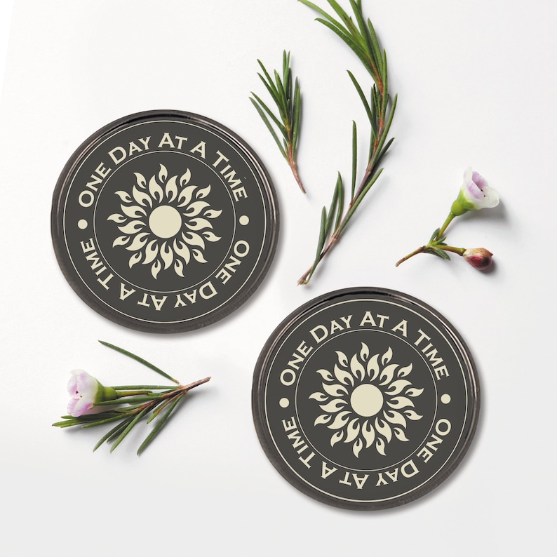 May include: Two black round coasters with a white sun design and the text "One Day At A Time" repeated around the sun. The coasters are on a white surface.