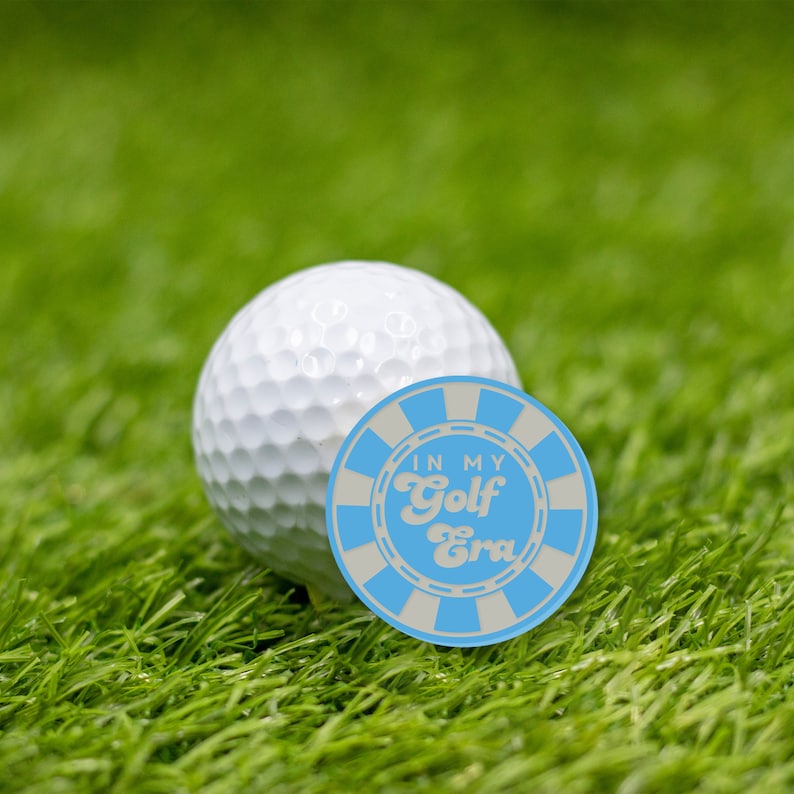 May include: A white golf ball with a blue and white circular sticker that says "In My Golf Era" in white text.