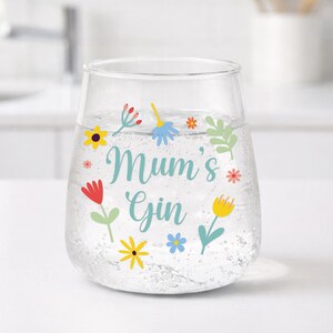 Może przedstawiać: Przezroczysty szklany kieliszek do ginu bez n&oacute;żki, wypełniony gazowanym płynem. Ozdobiony kolorowymi kwiatowymi ilustracjami i napisem "Mum's Gin" w kolorze turkusowym. Kwiaty są czerwone, ż&oacute;łte, niebieskie i r&oacute;żowe.