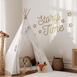 May include: A white teepee tent with a white pom-pom trim and a deer pillow inside. The teepee is set up in a room with a white wall that has gold star and "Story Time" decals.