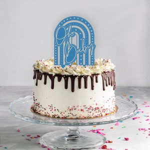 May include: A white cake with chocolate frosting and sprinkles topped with a blue 'Oh Baby' rainbow cake topper.
