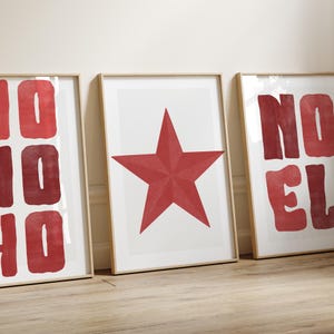 May include: Three framed art prints with a minimalist design. The prints feature red text and a red star on a white background. The frames are a light wood color, and the artwork is displayed on a wooden floor.