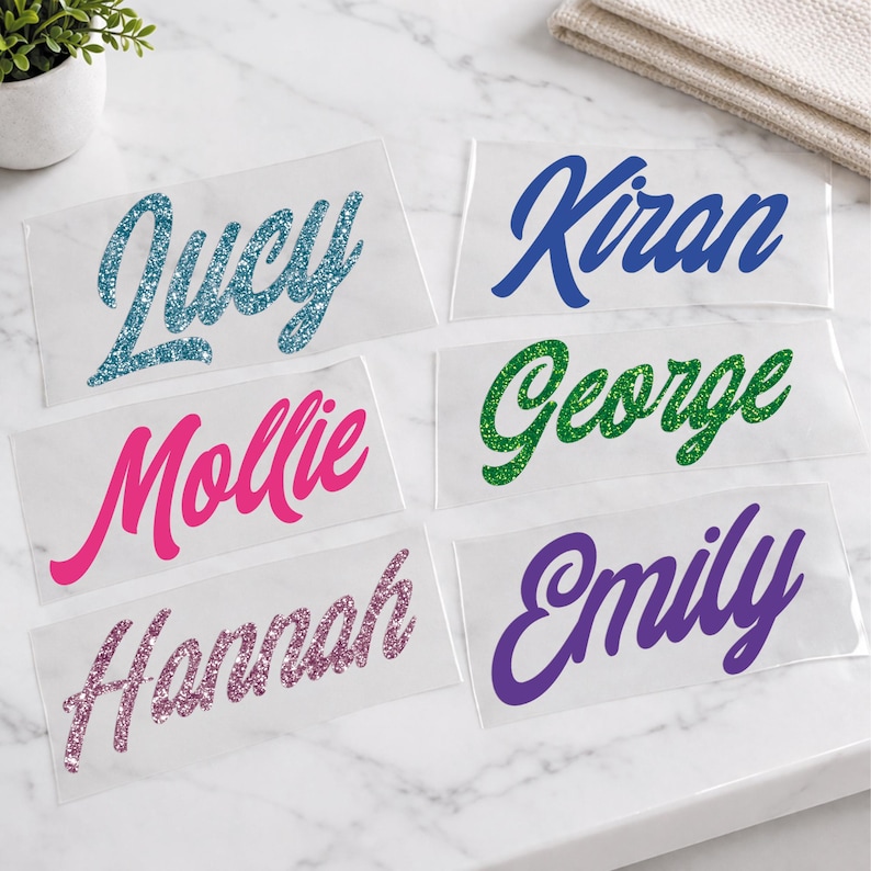 May include: Clear adhesive name decals in various colors, including blue, pink, green, purple, and glittery pink. Names include Lucy, Kiran, Mollie, George, Hannah, and Emily. Perfect for personalizing items.