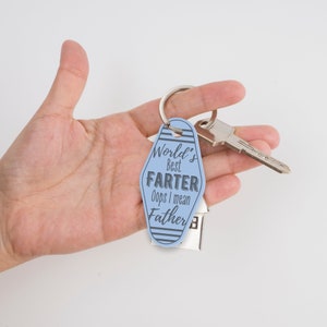 以下が含まれることがあります： 黒い文字で「World's Best Farter Oops I mean Father」と書かれた青いモテルのキーホルダー