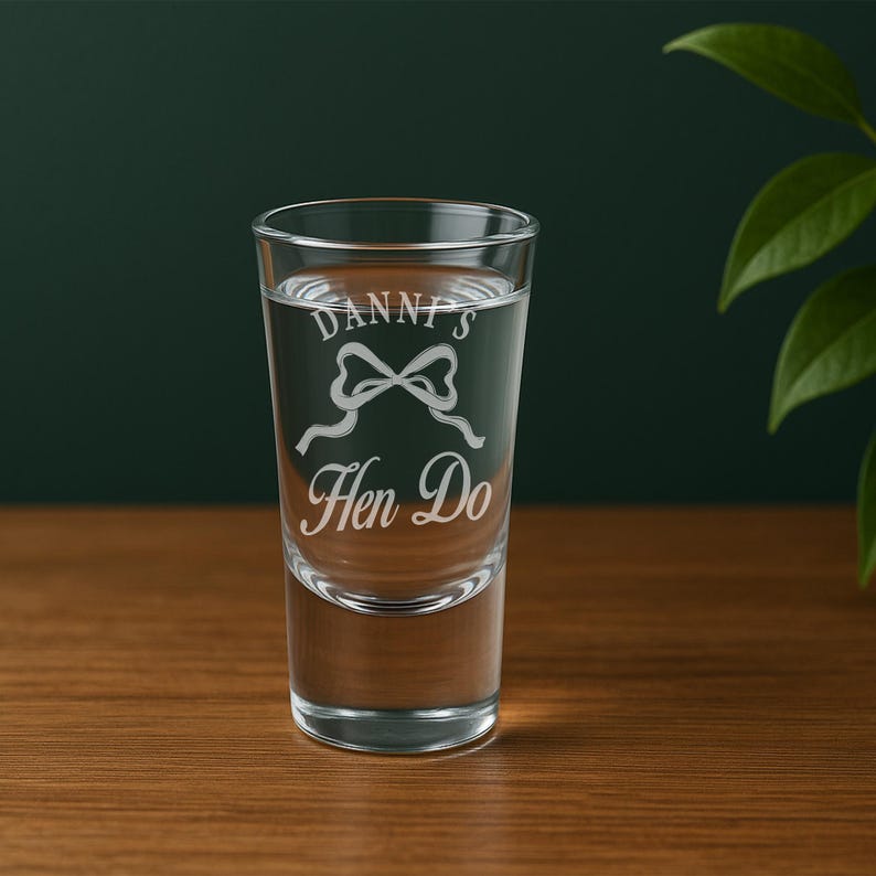 May include: A clear shot glass filled with liquid, featuring the text "Danni's Hen Do" in an elegant script, along with a bow graphic. The glass is set against a dark green background.