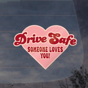 May include: A pink heart-shaped sticker with the words "Drive Safe" in red script above the text "Someone Loves You!" in a smaller, red font. The sticker is likely for a car window.