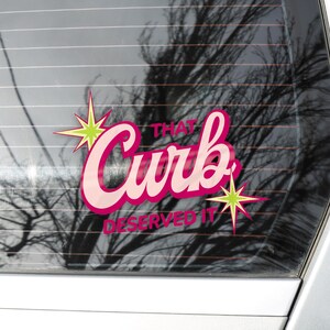 May include: A pink and white sticker with the words "That Curb Deserved It" in a retro font. The sticker has a starburst design in green and yellow. The sticker is on a car window.