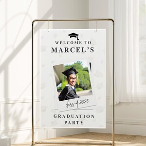 May include: A graduation party sign with the text "Welcome to Marcel's" and "Graduation Party". A photo of a graduate in a cap and gown is featured. The sign is suspended from a gold metal frame.