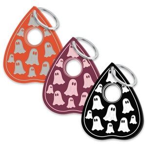 May include: Three different colored keychains shaped like a planchette, a tool used in a Ouija board game. The keychains are decorated with a pattern of white ghosts on a red, purple, and black background.