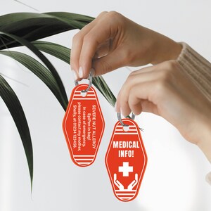 Pode incluir: Dois chaveiros de motel vermelhos com texto branco. Um chaveiro diz "SEVERE NUT ALLERGY EpiPen in bag! In case of emergency, please contact my mother, Shelly, at 01234 123456." O outro chaveiro diz "MEDICAL INFO!" com um sinal de mais branco e duas m&atilde;os segurando o sinal de mais.