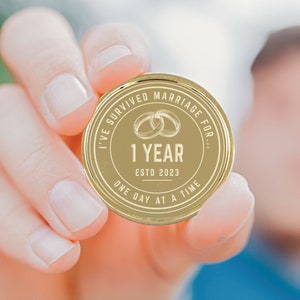 Puede incluir: Una moneda conmemorativa dorada con el texto "I've survived marriage for..." y "1 YEAR ESTD 2023 ONE DAY AT A TIME." Dos anillos entrelazados est&aacute;n en el centro. La moneda se sostiene en una mano.