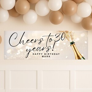 以下が含まれることがあります： 「Cheers to 30 years! Happy Birthday Mark」の文字とシャンパンボトルの描かれた誕生日バナー。背景は白で、ボケ効果があります。白とベージュの風船が画像上部に配置されています。