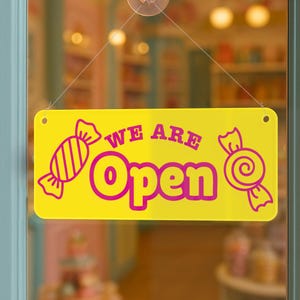 Puede incluir: Un letrero rectangular amarillo brillante con las palabras "WE ARE Open" en rosa. El letrero tiene gr&aacute;ficos de caramelos rosas a cada lado. El letrero est&aacute; colgado en una puerta de cristal.