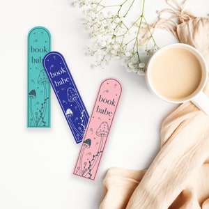 May include: Three colorful bookmarks with a mushroom and floral design. The bookmarks are blue, pink, and teal and each has the text "book babe" printed on them.