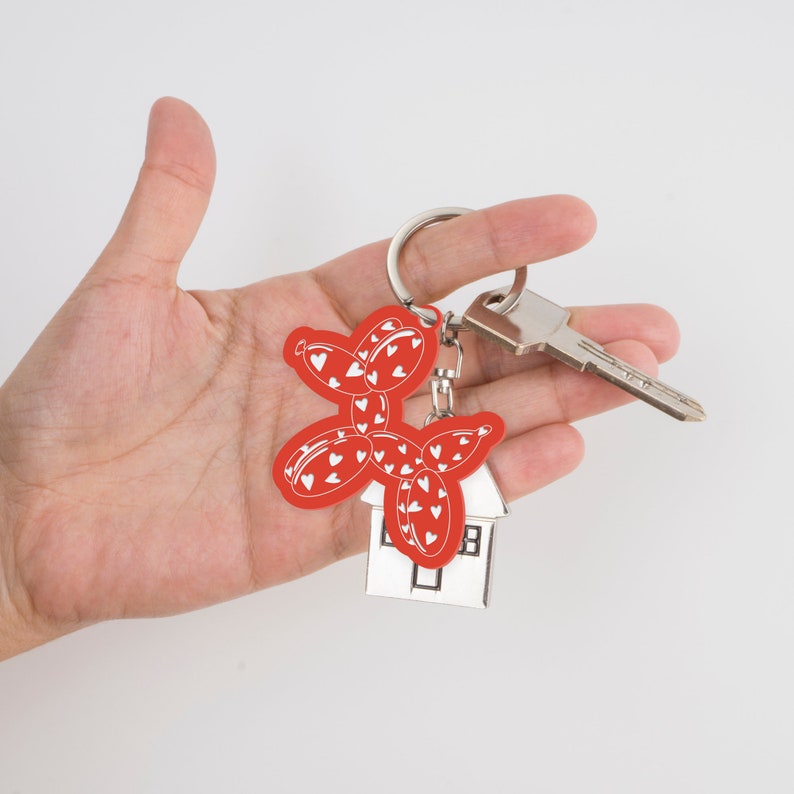 May include: A red balloon dog keychain with white heart details, attached to a silver keyring with a house charm and a key. The keychain is held in a hand against a white background.