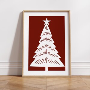 May include: A framed print featuring a white Christmas tree design on a deep red background. The tree is topped with a star and has a minimalist style. The frame is a light wood color, and the print is displayed on a wooden floor.